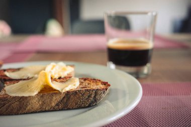 Bir fincan kahvenin yanında bir tabakta sunulan kızarmış ekmek ve tereyağlı basit kahvaltı sahnesi. Sıcak sabah atmosferi günlük yaşam tarzını ve rutinini temsil ediyor..