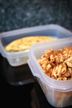 Plastik bir yiyecek kabında saklanan ve arka planda hazırlanmış omlet ile birlikte domates soslu ev yapımı makarna. Yemek hazırlama, yemek pişirme ve ev yemeği konsepti..