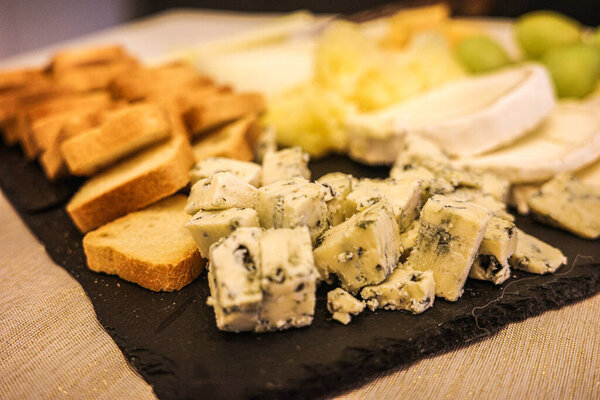 Assorted cheese platter with blue cheese pieces and slices of bread served on a slate board. Close-up food photography with shallow depth of field.