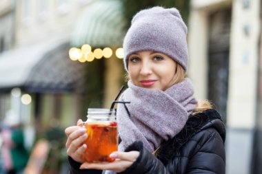 Eşarplı ve şapkalı genç bir kız sokakta sıcak bir içki içiyor. Elinde sıcak çayla Noel sokaklarında yürü. Noel indirimi.