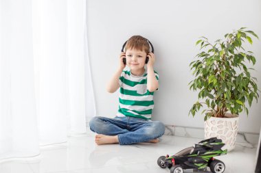 Küçük bir çocuk, parlak bir odada yerde oturmuş oyuncak bir arabanın yanında kulaklıkla müzik dinliyor..