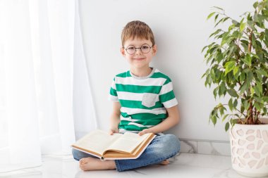 Yeşil çizgili tişörtlü bir çocuk yerde oturmuş kitap okuyor, boş zamanlarını yeşil bir ağacın yanında ders çalışarak geçiriyor..