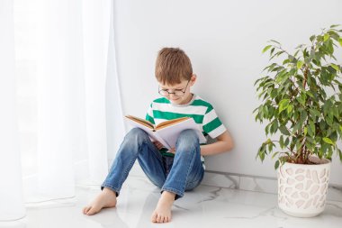 Yeşil çizgili tişörtlü bir çocuk yerde oturmuş kitap okuyor, boş zamanlarını yeşil bir ağacın yanında ders çalışarak geçiriyor..