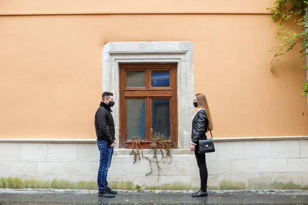 Two people, a guy and a girl in black clothes and a medical mask, reach out to each other, social distance, a date in a pandemic. In the background, the window of an old yellow house