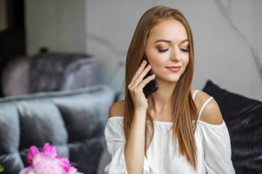 Evdeki deri koltukta oturan genç kızıl saçlı kadın cep telefonuyla konuşuyor. Gülümseyen güzel Avrupalı kız elinde kağıt bardak tutuyor. İçinde şakayık çiçekleri olan modern bir daire.