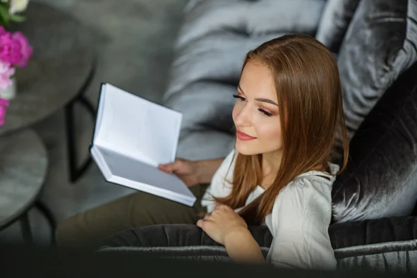 自宅でベロアソファに座りながら本を持ち考えながら読書をしている若い赤毛の女性 白いブラウスとパンツを着た笑顔の美しいヨーロッパの女の子のオーバーヘッドビュー 現代のアパートのインテリア — ストック写真