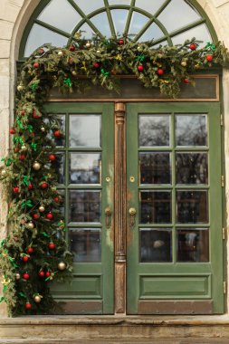 Old green wooden door adorned with Christmas garland, red and gold ornaments, and festive greenery in a holiday street setting.