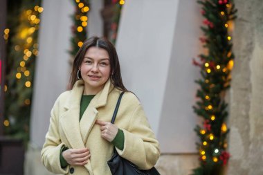 Yün ceketli genç bir kadın, arka planda Noel ışıkları ve çelenklerle süslenmiş bir şehir caddesinde hafifçe gülümsüyor..