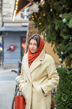 Bej ceket ve turuncu eşarp giyen kendine güvenen bir kadın kış tatilinde elinde kırmızı bir alışveriş çantasıyla şehir caddesinde yürüyor..