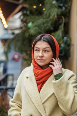 Sıcak bej bir ceket ve turuncu eşarp giyen kendine güvenen genç bir kadın, şehrin şenlikli bir caddesinde süslü bir Noel ağacının yanında duruyor..