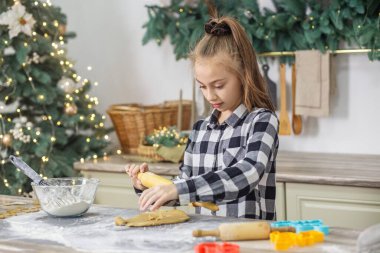 Genç kız, Noel kurabiyeleri için ağaç ışıkları ve şenlikli aletlerle donatılmış bir Noel mutfağında hamur topluyor..