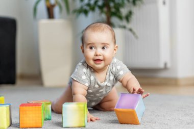 Puantiyeli mutlu bebek bebek tulumu halıda sürünürken rahat bir evde yumuşak renkli köpük bloklarıyla oynuyor..