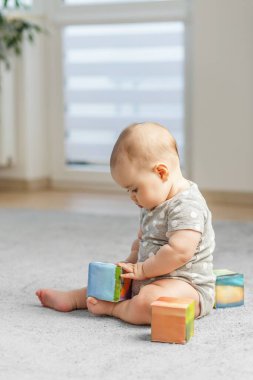 Puantiyeli bebek tulumu giymiş konsantre bir şekilde halıya oturur ve evdeki yumuşak renkli bloklarla sessizce oynar..