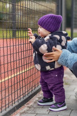 Sıcak giysili meraklı bebek soğuk bir günde soğuk bir kardeşin nazik desteğiyle metal bir çitin yanında duruyor..