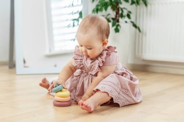 Pembe elbiseli şirin bir kız, ahşap zeminde oturuyor ve ev ortamında renkli yüzüklerle oynuyor..