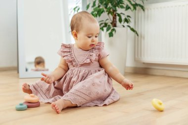 Pembe elbiseli bebek ahşap zeminde oturuyor, renkli bir yüzüğe uzanıyor, erken oyun ve motor geliştirme ile meşgul oluyor..