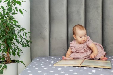 Pembe elbiseli bebek yıldız desenli bir yatakta tek başına oturuyor, sakin, rahat bir odada renkli bir resim kitabına bakıyor..