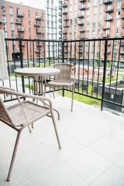 Hasır sandalyeli ve masalı modern şehir dairesi balkonu, konut binalarına bakan ve aşağıdaki ortak avlusu.