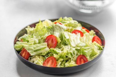 Açık arkaplanda siyah bir kasede taze marul ve kiraz domatesinden yapılmış sağlıklı Sezar tarzı salatanın yakın çekimi..