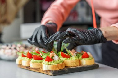 Avokado sosu, karides, domates dilimleri ve taze yeşilliklerle kaplı baget kanapelerini dikkatlice birleştirirken siyah eldivenler.