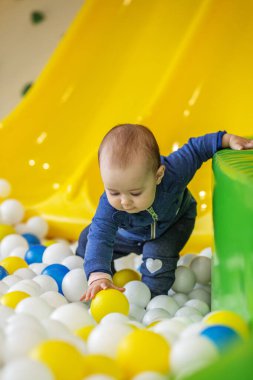 Mavi elbiseli bir bebek, kapalı alandaki renkli bir top havuzunda oynuyor, sarı plastik bir top kapmak için uzanıyor..