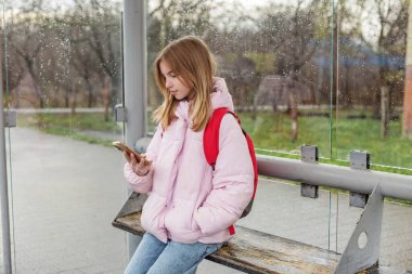 Pembe ceketli ve kırmızı sırt çantalı genç bir kız yağmurlu bir günde ıslak bir otobüs durağında oturmuş akıllı telefonuna bakıyor..