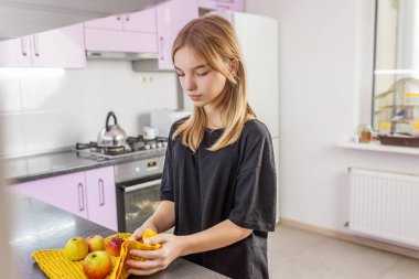 Siyah gömlekli genç bir kız modern bir mutfakta sarı havluyla elma kurutuyor. Sağlıklı yemek rutininin bir parçası..