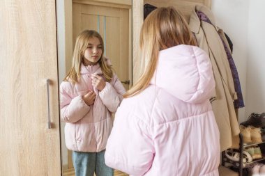 Pembe bir kabarık ceket giyen genç bir kız koridor aynasında kendine bakarken fermuarını çekip dışarı çıkmaya hazırlanıyor..