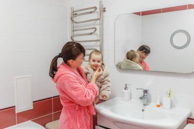 Bornozlu sevecen anne, bebeğine nazikçe bakar. Lavabonun yanındaki parlak, modern banyoda, aynaya yansıyan..