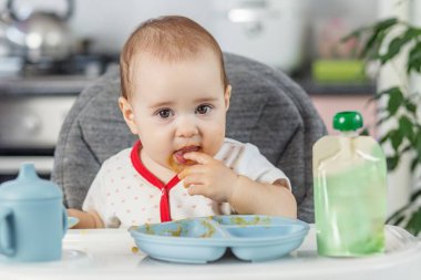 Kendi kendini besleyen bir bebeğin yakın çekimi, mutfakta dağınık yemek saatlerinde yüksek sandalyedeki bir tabaktan püre yemek..