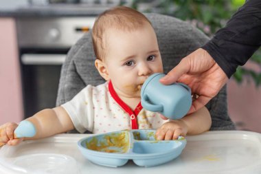Bebek, ağzında yemekle yüksek sandalyede oturuyor. Yetişkinler silikon bardaktan içerken kaşık tutuyor..