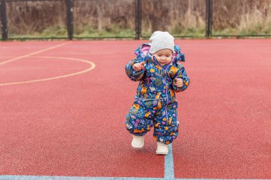 Renkli bir kış elbisesi giymiş sevimli çocuk soğuk bir günde kırmızı açık hava spor sahasında tek başına yürür..