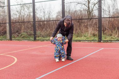 Kadınlar, her ikisi de kışlık giysiler içinde sıcak hava mevsiminde açık hava spor sahasında yürümeyi öğrenen bebeklere destek oluyor..