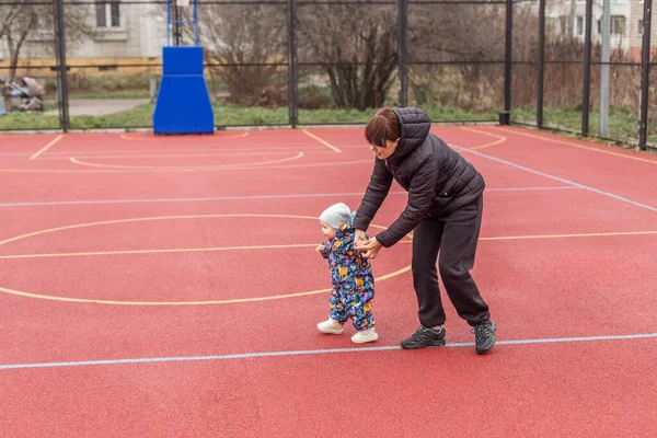 Anne, bebeğine kışın açık hava basketbol sahasında adım atmasında yardım eder. İkisi de mevsimlik kıyafetler giyer..