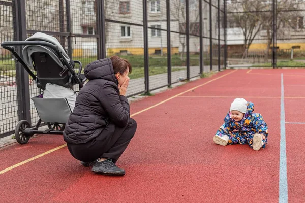 Renkli kar tulumuyla gezen çocuk düştükten sonra oyun sahasında otururken, anne şefkatli ve keyifli bir ifadeyle izliyor..