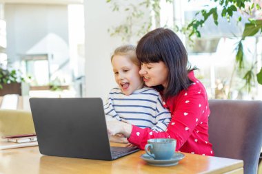 Gülümseyen anne ve küçük kızı bir masada birlikte oturuyorlar, mutlu bir şekilde kapalı bir ortamda dizüstü bilgisayara bakıyorlar..
