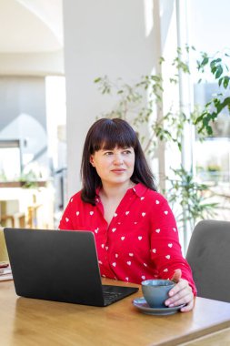 Kırmızı kalp desenli gömlekli bir kadın bir masada dizüstü bilgisayar ve kahveyle oturur, güneşli bir ofis alanına bakar..