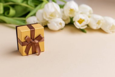 Bej bir yüzeye yerleştirilmiş bir buket beyaz lalenin yanına yerleştirilmiş kahverengi kurdeleli küçük altın hediye kutusu kutlama ve minnettarlığı simgeliyor..