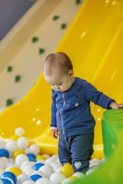 Young baby standing still and looking down in a colorful ball pit area with yellow slide and green padded wall nearby.
