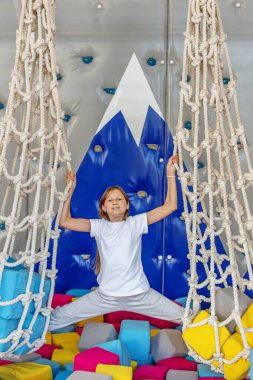 Genç bir kız renkli bir oyun bahçesinde köpük bloklar ve tırmanma duvarı arka planıyla iki halat arasında dengede duruyor..