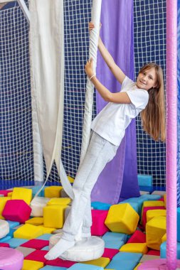 Yumuşak köpük bloklarıyla dolu renkli bir oyun merkezinde tırmanan ve sallanan mutlu genç kız..