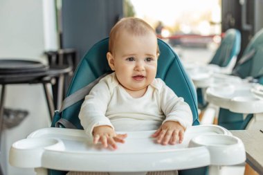 Neşeli bebek yüksek bir sandalyede oturuyor, etrafı aydınlık bir mekanla meşgul ediyor, muhtemelen yemek ya da oyun için bekliyor..