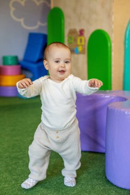 Sevimli bebek ilk bağımsız adımlarını atıyor. Kolları açık ve dengeli. Renkli, güvenli bir kreş ortamında yumuşak oynanıyor..