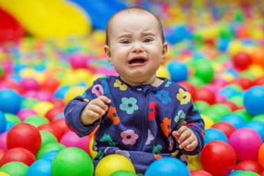 Üzgün bebek, kapalı bir top havuzunda renkli plastik topların arasında otururken, parlak çiçekli bir tulum giyerken ağlıyor..