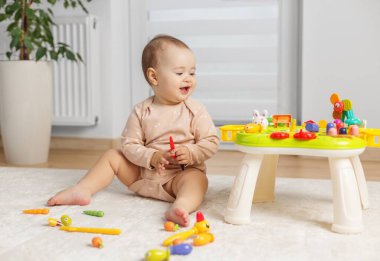 Gülümseyen bebek yumuşak bir halının üzerinde otururken renkli eğitici oyuncaklardan hoşlanır, evde öğrenmeyi ve motor becerileri geliştirmeyi teşvik eder.