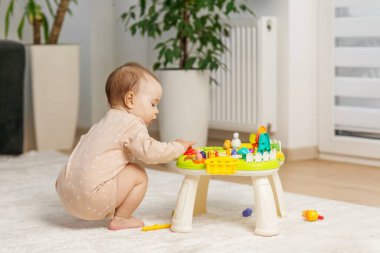 Etkileşimli oyuncak masasının yanına çömelmiş meraklı bebekler, derinden renkli parçalara odaklanmış, koordinasyonu arttırıyor ve evde erken öğrenimi geliştiriyor..