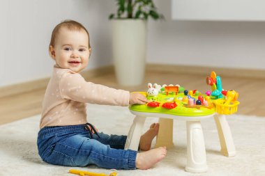 Mutlu bebek yumuşak bir halıya oturur, rahat, çocuk dostu bir evde, parlak etkileşimli bir oyuncak masasıyla oynar..