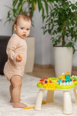 Yumuşak vücutlu meraklı bir bebek, rahat bir evin içinde halı döşeli bir zeminde dururken eğitici bir oyuncak masasını keşfediyor..