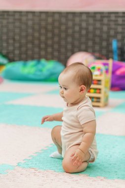 Renkli kreş ortamında oyuncaklarla ve eğitim faaliyet merkezleriyle yumuşak köpük minderinde otururken denge uygulayan küçük bir bebek..