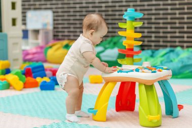 Bebek renkli aktivite masasında duruyor, güvenli ve uyarıcı bir kreşte koordinasyon becerileri geliştiriyor ve oynuyor..